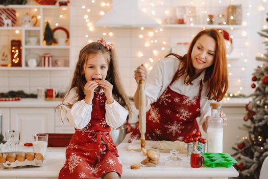 A little girl in the New Year's kitchen is sitting on the table with cookies in her hands, and her mother is preparing dough.