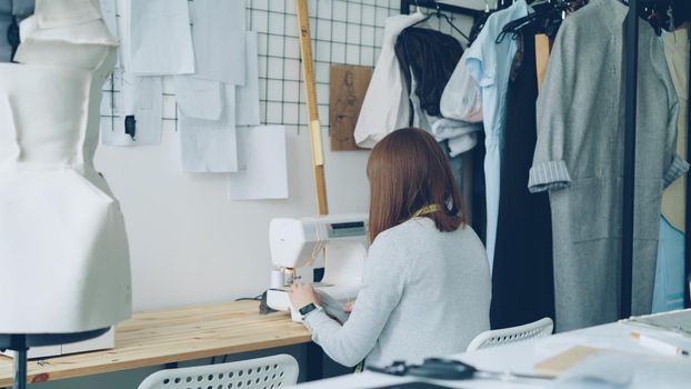 Back view of young woman clothing designer working with sewing machine sitting at studio desk. Sewing items, tailoring dummy and half-finished women's clothes are visible.