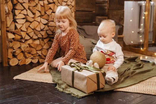 Little brother and sister play on Christmas eve in a beautiful house decorated for the New Year holidays. Children are playing with a Christmas gift. Scandinavian-style interior with live fir trees and a wooden staircase.
