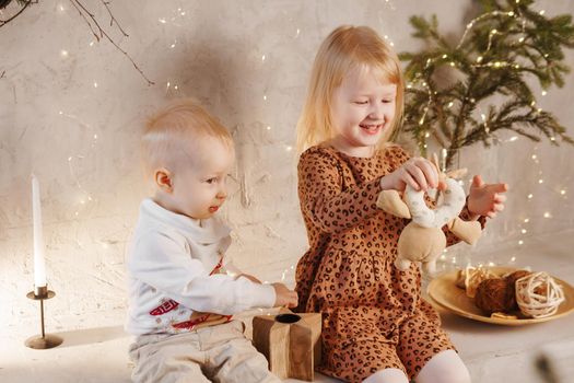Little brother and sister play on Christmas eve in a beautiful house decorated for the New Year holidays. Children are playing with a Christmas gift. Scandinavian-style interior with live fir trees and a wooden staircase.