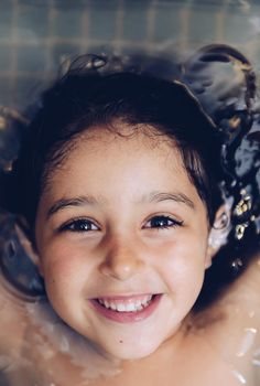 vertical portrait of a happy pretty little girl smiling lying in the tub while taking a bath in the bathtub, kids hygiene concept, copy space for text