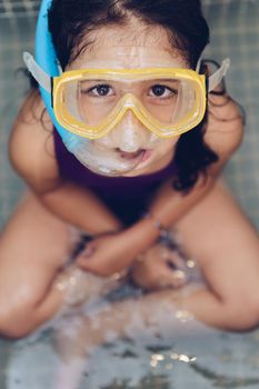 vertical portrait of a serious beautiful little girl sitting with snorkel goggles in the tub while taking a bath in the bathtub, kids hygiene concept, copy space for text