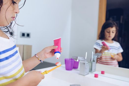 little girl putting toothpaste on the brush, after taking a shower she prepares to brush her teeth in front of the mirror of the bathroom, kids hygiene concept