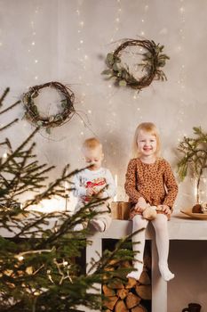 Little brother and sister play on Christmas eve in a beautiful house decorated for the New Year holidays. Children are playing with a Christmas gift. Scandinavian-style interior with live fir trees and a wooden staircase.