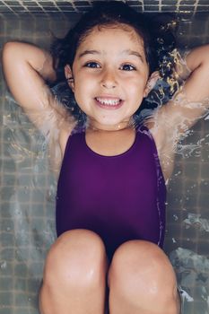 vertical photo of a beautiful little girl having fun and smiling lying in the tub while taking a bath in the bathtub, kids hygiene concept
