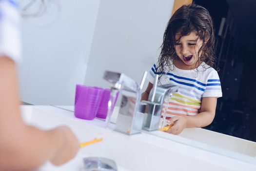 funny little girl wetting the toothbrush, after taking a shower she prepares to brush her teeth in front of the mirror of the bathroom, kids hygiene concept