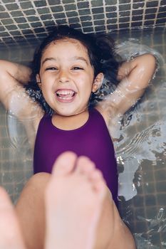 vertical photo of a playful beautiful little girl having fun and laughing lying in the tub and showing the feet while taking a bath in the bathtub, kids hygiene concept
