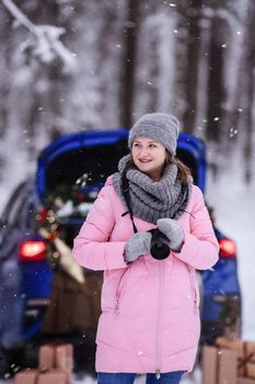 A woman in a winter snow-covered forest in the trunk of a car decorated with Christmas decor. A female photographer holds a camera in her hands.