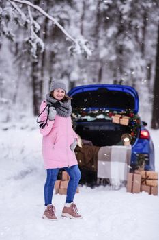 A woman in a winter snow-covered forest in the trunk of a car decorated with Christmas decor. A female photographer holds a camera in her hands.