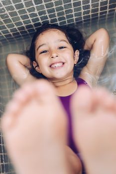 vertical photo of a playful beautiful little girl having fun and smiling lying in the tub and showing the feet while taking a bath in the bathtub, kids hygiene concept