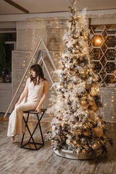 A brunette woman in a beautiful light tight dress next to a Christmas tree. The magical atmosphere of Christmas Eve on December 25. Decoration for the holiday in a light style with lots of lights and light bulbs.