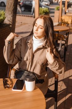 A stylish brunette woman is sitting at a table in an outdoor cafe with coffee and a phone. Autumn walk.