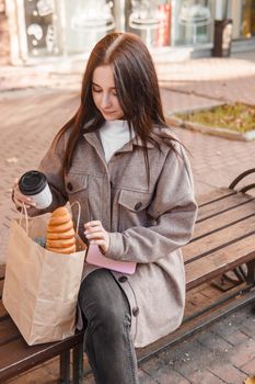A stylish brunette woman walks around the autumn city. The brunette is sitting on a bench outside with a glass of coffee and a phone, holding a craft bag with a baguette.