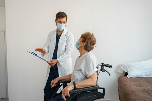 Doctor in mask explaining to a sick patient in wheelchair details of treatment. High quality photo