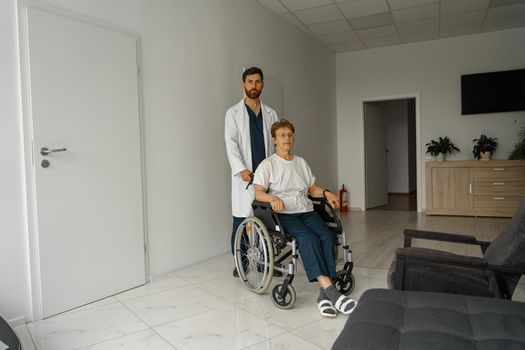 Professional male doctor carrying patient on wheelchair in medicine clinic hall. High quality photo