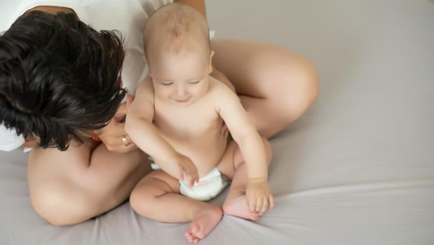 Mother and child on a white bed. Mom and baby boy in diaper playing in bedroom. Parent and little kid relaxing at home. Family having fun together