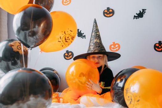 Children's Halloween - a girl in a witch hat and a carnival costume with airy orange and black balloons at home. Ready to celebrate Halloween.