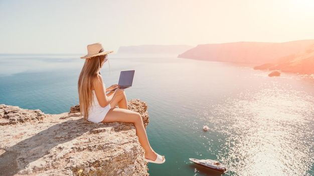 Successful business woman in yellow hat working on laptop by the sea. Pretty lady typing on computer at summer day outdoors. Freelance, travel and holidays concept.