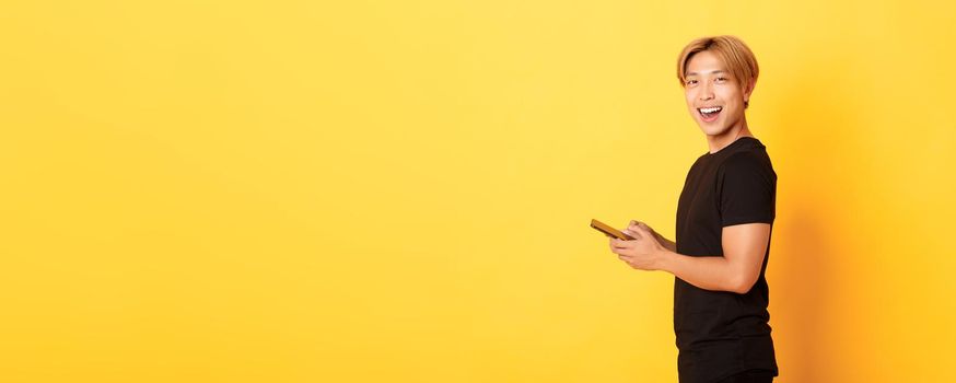 Portrait of handsome stylish asian guy in black outfit, using mobile phone and turning head at camera with satisfied smile, yellow background.