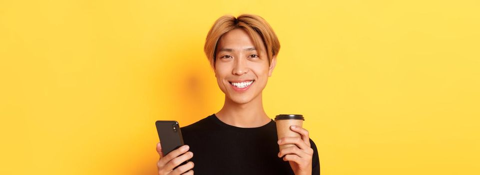 Close-up of handsome young asian guy smiling happy, using smartphone and drinking takeaway coffee, standing over yellow background.