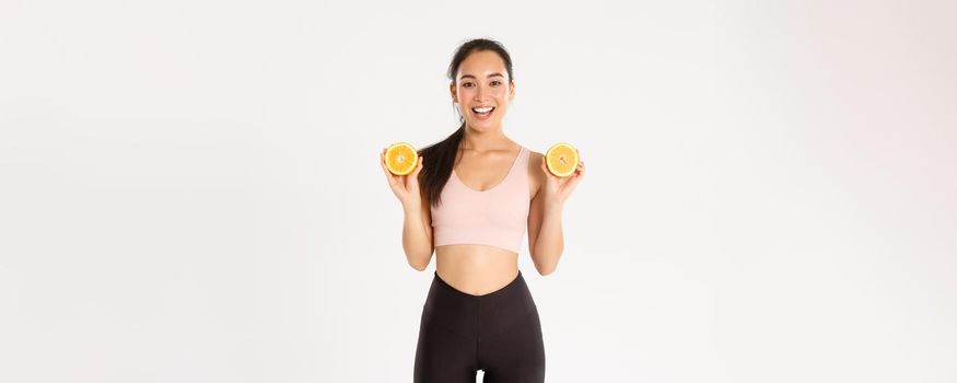 Sport, wellbeing and active lifestyle concept. Portrait of smiling healthy and slim asian girl advice eating healthy food for breakfast, gain energy for workout, hold two halves of orange.