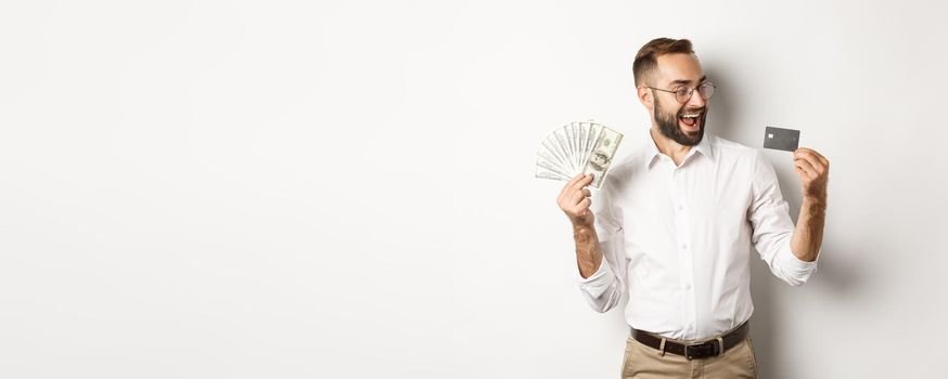 Excited businessman holding money and looking at credit card, standing over white background.