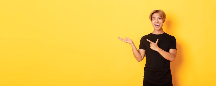 Portrait of handsome smiling asian guy holding something on hand and pointing finger at it, standing yellow background pleased.