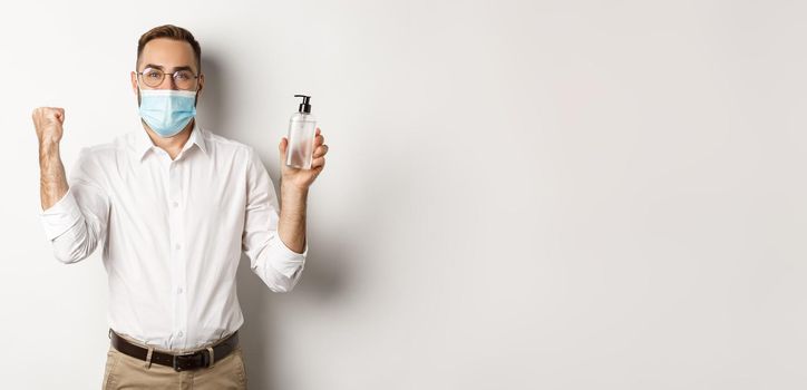 Covid-19, social distancing and quarantine concept. Cheerful manager in medical mask showing hand sanitizer, standing over white background.