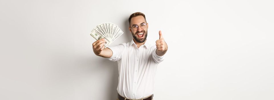 Excited man showing thumbs up and money, earning cash, standing over white background.