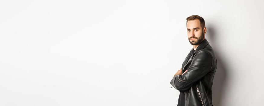 Profile of confident and handsome bearded man in black leather jacket, turn face at camera and looking serious, white background.