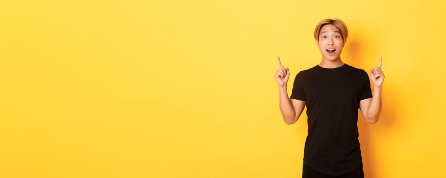 Portrait of surprised and excited asian guy looking amused, pointing fingers up, yellow background.