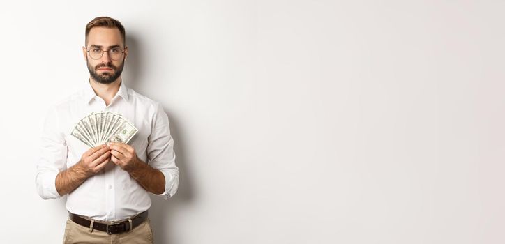 Serious businessman holding money, showing dollars, standing over white background.