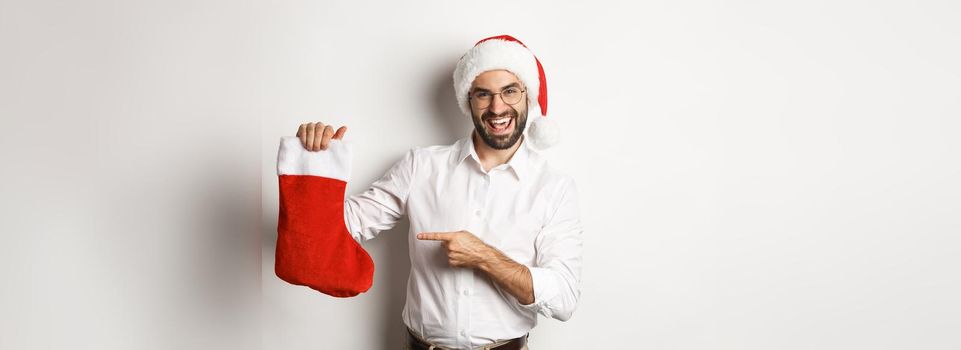 Happy man in santa hat celebrating winter holidays, pointing at christmas sock and smiling, holding gifts, standing over white background.