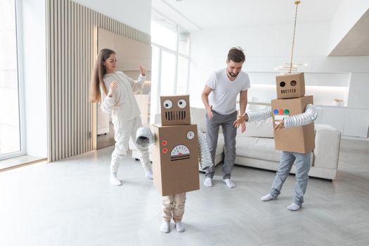 Happy family of parents and two children playing dancing like robots at home, children wearing handmade moving box costume of cardboard
