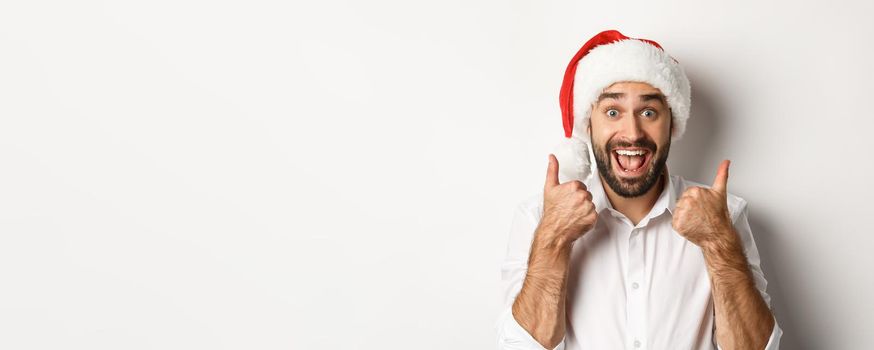 Party, winter holidays and celebration concept. Man enjoying christmas, wearing santa hat and showing thumb up with excited face, white background.