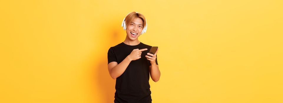Joyful satisfied asian handsome guy, listening music or good podcast in headphones, pointing finger at smartphone with pleased smile, yellow background.