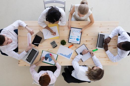 Top view of business people in formal wear sitting in boardroom at desk and analyzing data. On desk are laptops, graphs, tablets and paperwork.