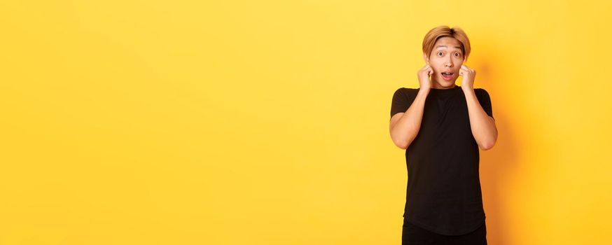 Excited handsome blond guy in black clothes, looking tempted and standing over yellow background.