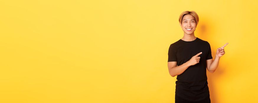 Portrait of satisfied smiling blond guy, asian student pointing fingers right, showing way, standing yellow background.