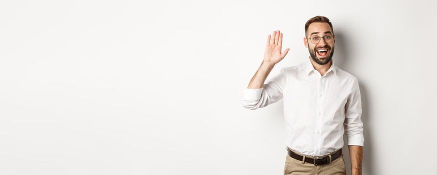 Friendly smiling man in glasses saying hello, waving hand in greeting, standing over white background.