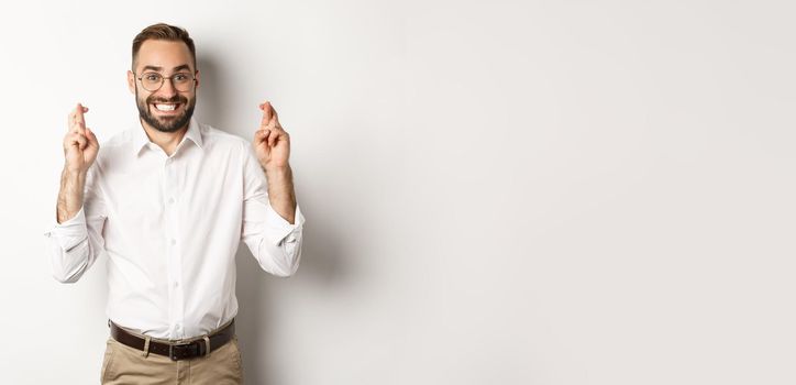 Hopeful businessman smiling and cross fingers for good luck, making a wish, standing over white background.