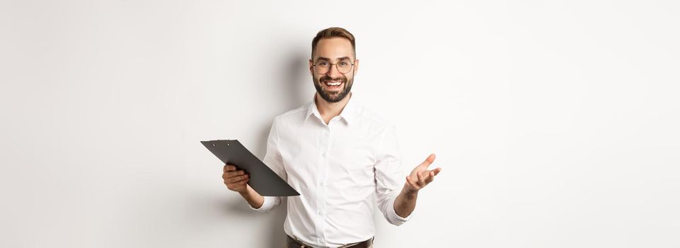 Handsome boss looking satisfied, holding clipboard and praising you, standing over white background.