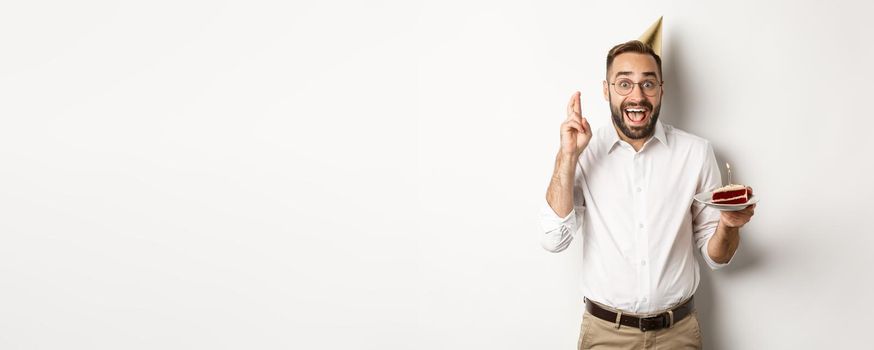 Holidays and celebration. Excited man having birthday party, making wish on b-day cake and cross fingers for good luck, standing against white background.