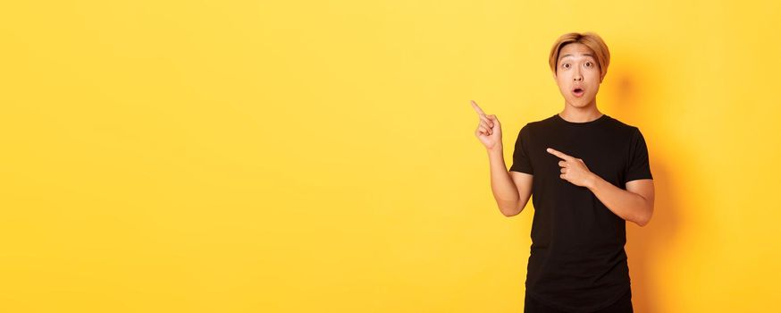 Portrait of impressed and excited handsome asian guy in black t-shirt, react to your logo, pointing fingers upper left corner.