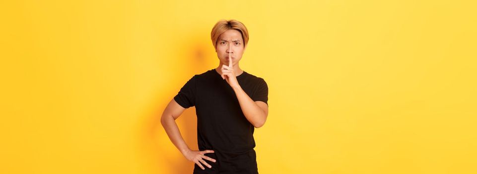 Portrait of angry asian man frowning, shushing at camera disappointed, standing yellow background.