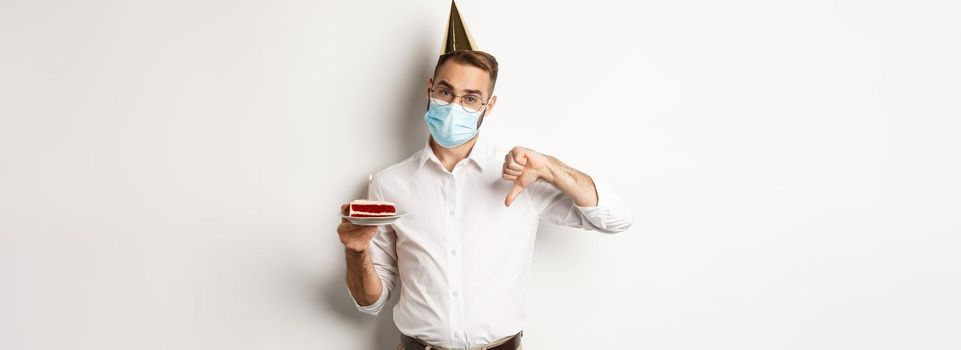 Coronavirus, quarantine and holidays. Man showing thumb down as disappointed with birthday party, wearing face mask and holding bday cake, white background.