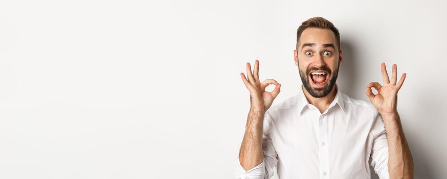 Close-up of amazed caucasian guy agreeing, showing okay signs and looking impressed, recommending product, white background.