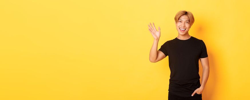 Portrait of handsome friendly asian guy in black outfit, waving hand to say hello and smiling, greeting someone, standing yellow background.