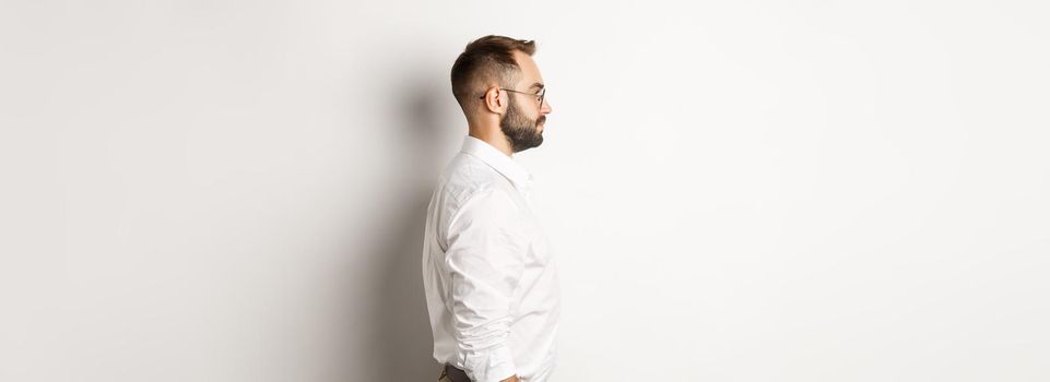 Profile of young businessman in white collar shirt and beige pants, looking left, standing against studio background.