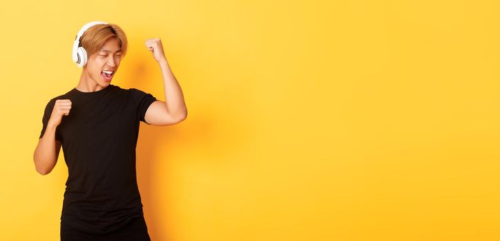 Carefree attractive asian guy with blond hair, singing along and dancing as listening to music in wireless headphones, standing yellow background.